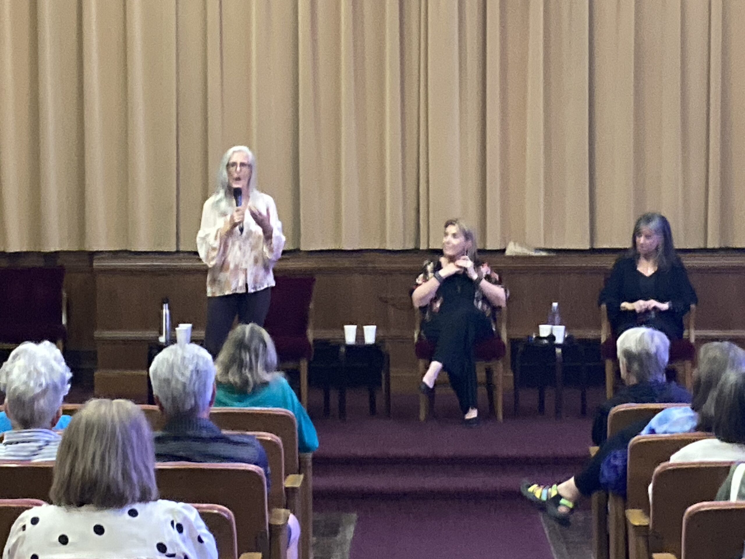 A speaker addresses an audience in a theater setting, with two women seated on stage