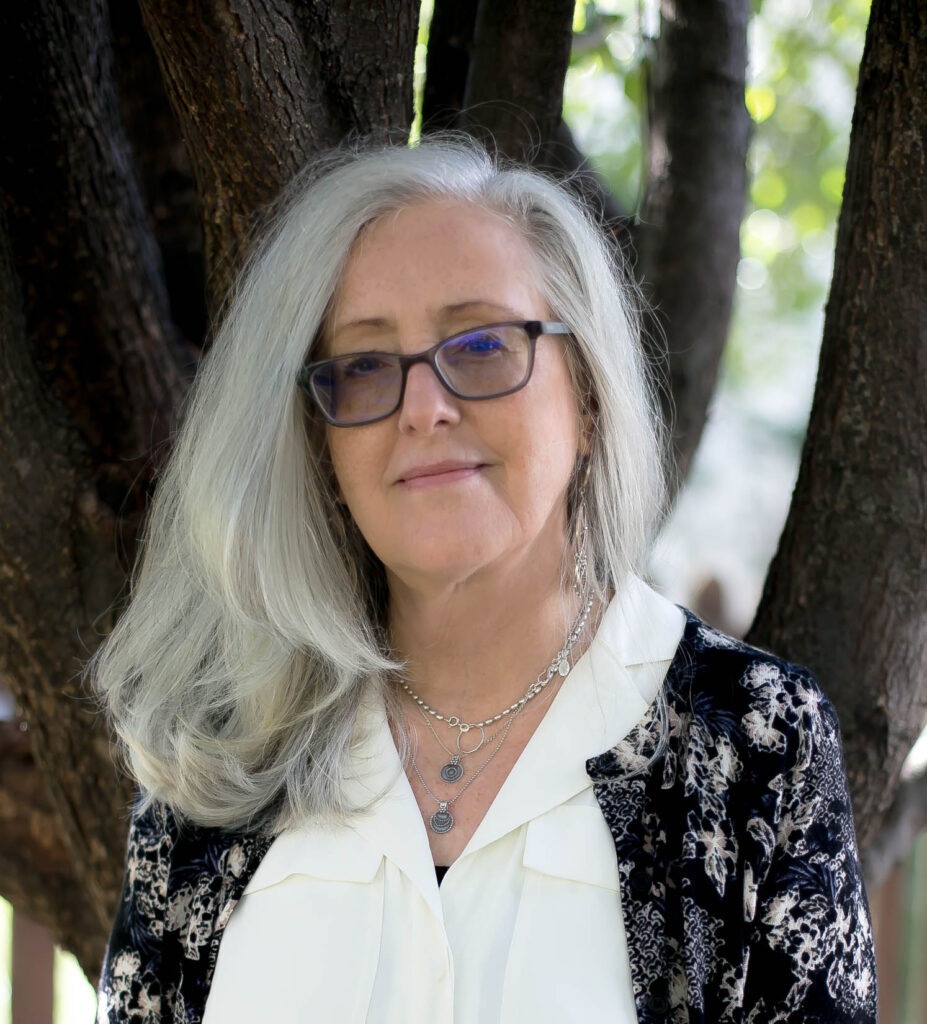 A woman with long silver hair and glasses stands outdoors